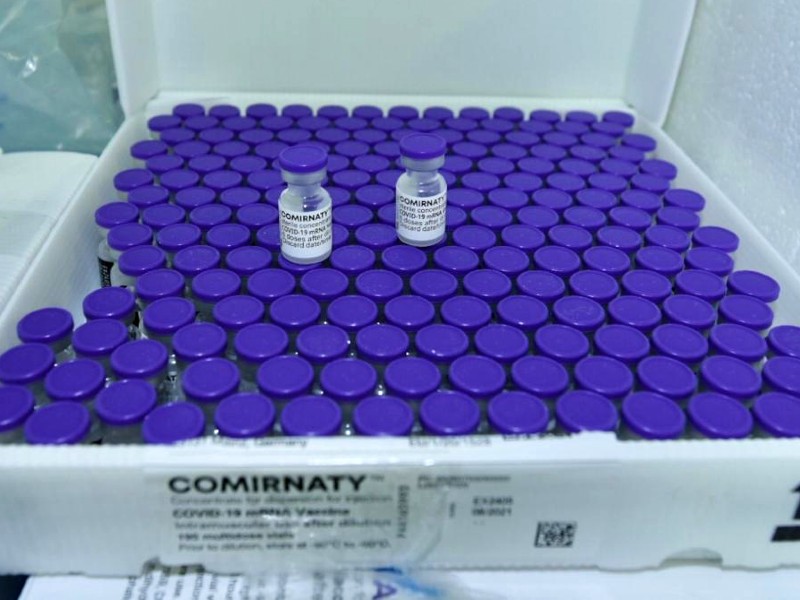 Foto das vacinas da Pfizer. Os frascos têm tampa azul e rótulo branco com o nome da vacina (Comirnaty) e estão dispostos em uma caixa branca. Duas ampolas estão em destaque em cima das demais